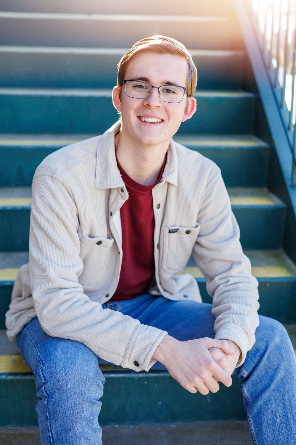 Smiling young man with glasses sitting on teal stairs wearing a cream jacket and red shirt.