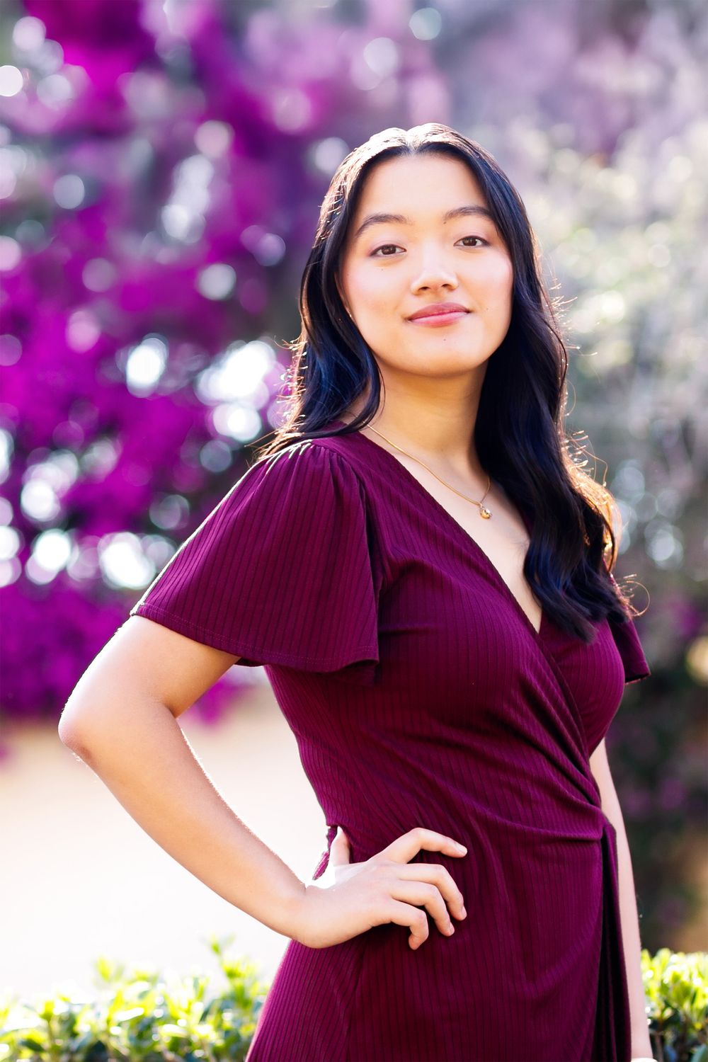 Young Asian woman in burgundy wrap dress poses confidently outdoors with purple bougainvillea flowers in background.