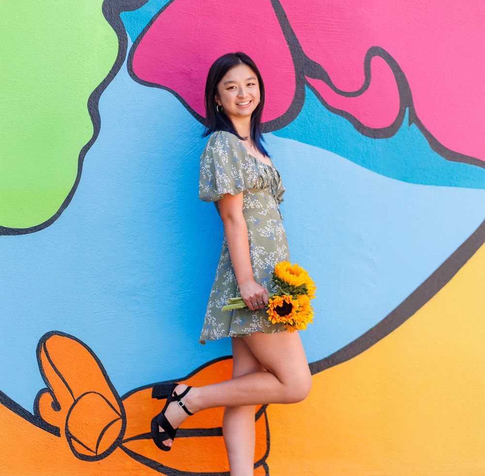 Young woman in floral dress holding sunflowers, posing against a vibrant colorful mural wall.