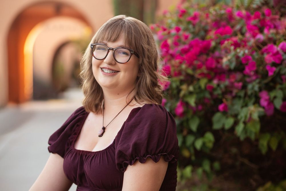 Young woman with glasses smiling outdoors near vibrant pink flowers and arched architecture in a maroon dress.