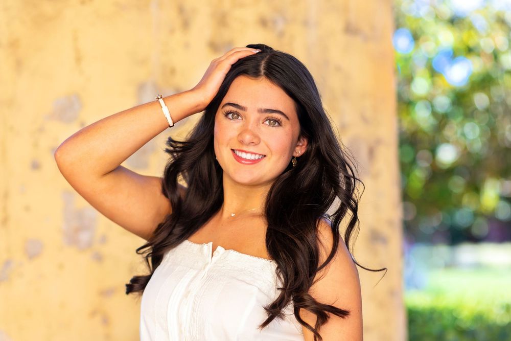 Smiling young woman with long dark hair in white top, posing outdoors with hand in hair.