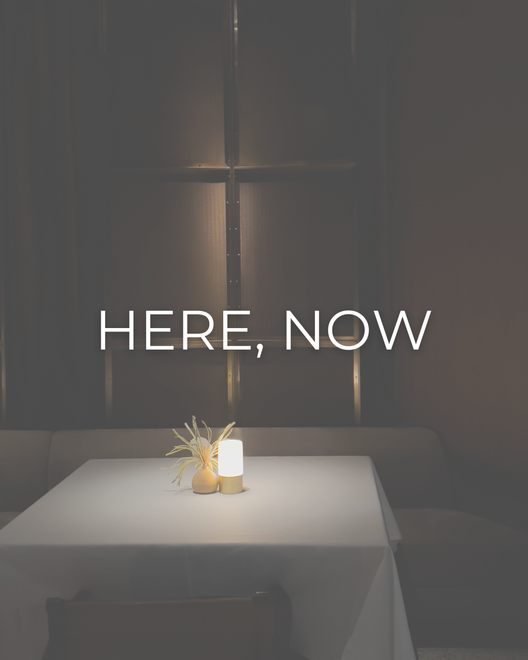A minimalist white table with a lit candle and small vase against dark walls, with text reading 'here, now' overlaid.