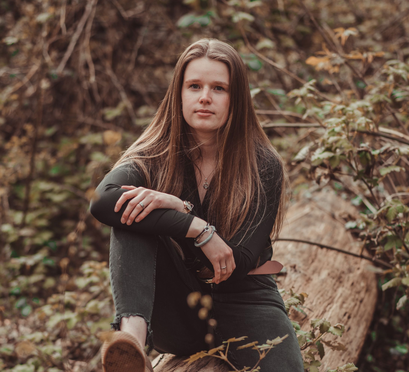 A person with long auburn hair poses gracefully among autumn foliage in a forest setting wearing dark clothing.