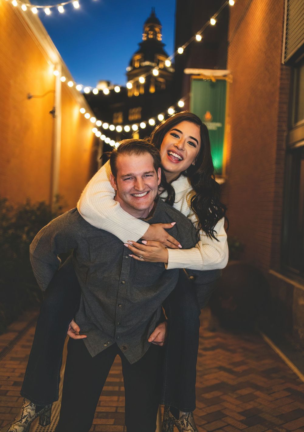 A joyful piggyback ride under string lights with a historic building dome in the background.