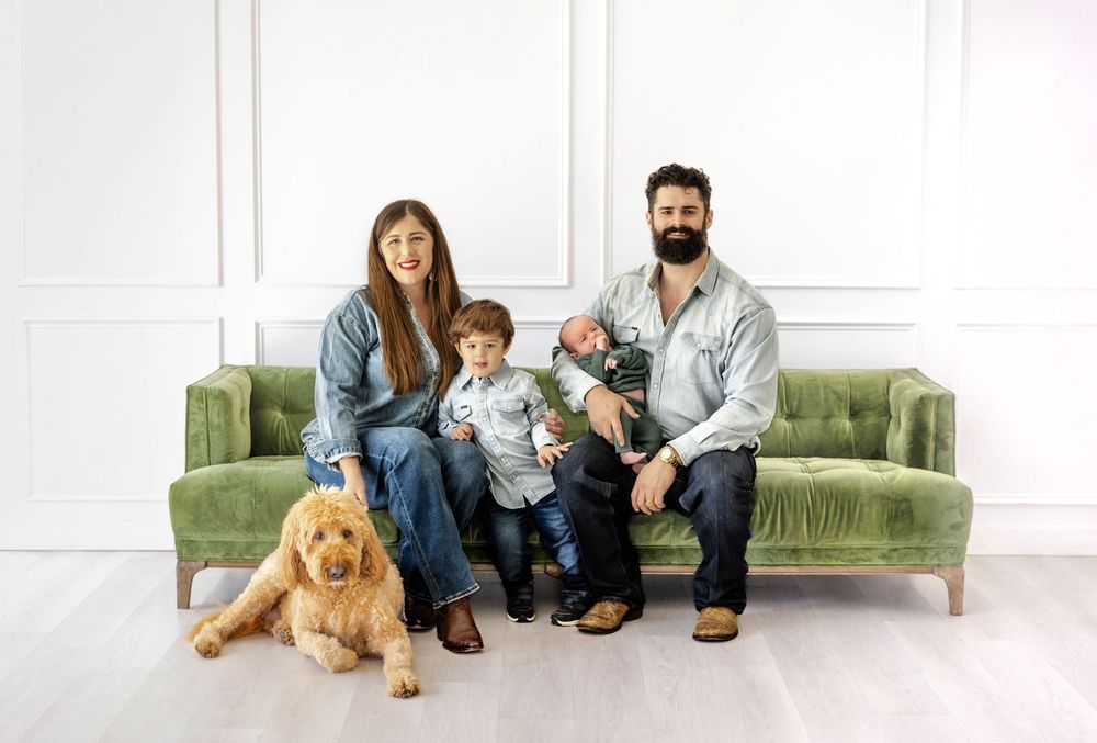 A family poses in a living room studio setting with their dog and a newborn baby.
