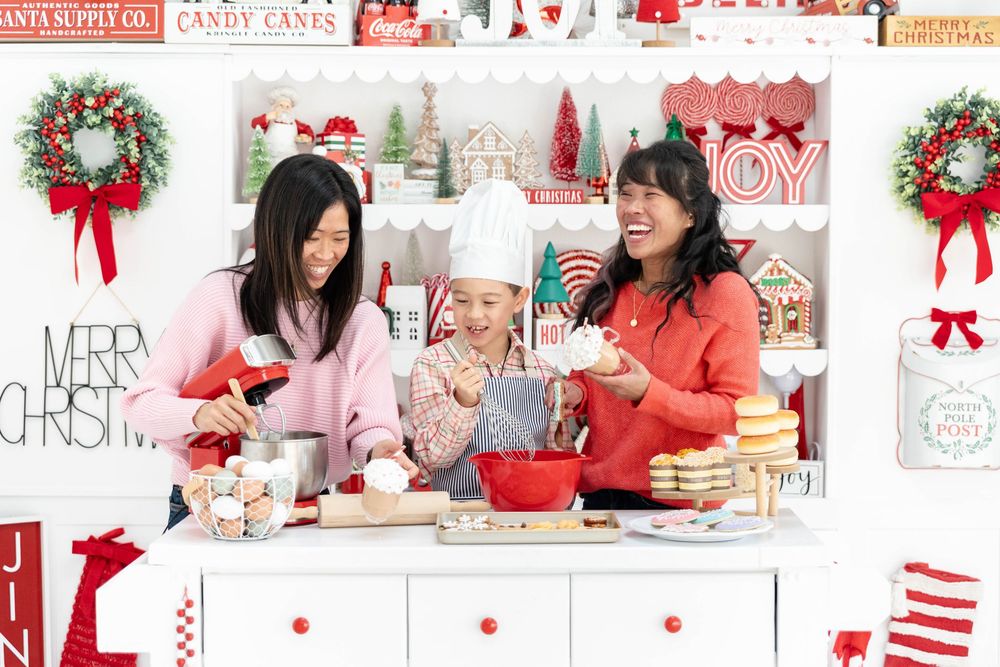 Two sisters with dark hair in a Christmas themed kitchen with one of their sons. The little boy is wearing a chefs hat.