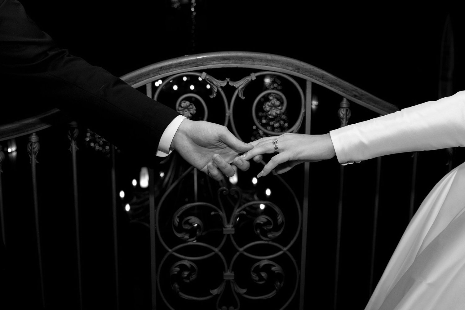 Black and white photo of hands reaching toward each other against ornate iron railing.