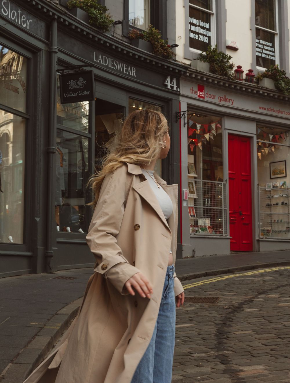 A person in a beige trench coat walks down a historic cobblestone street with storefronts.