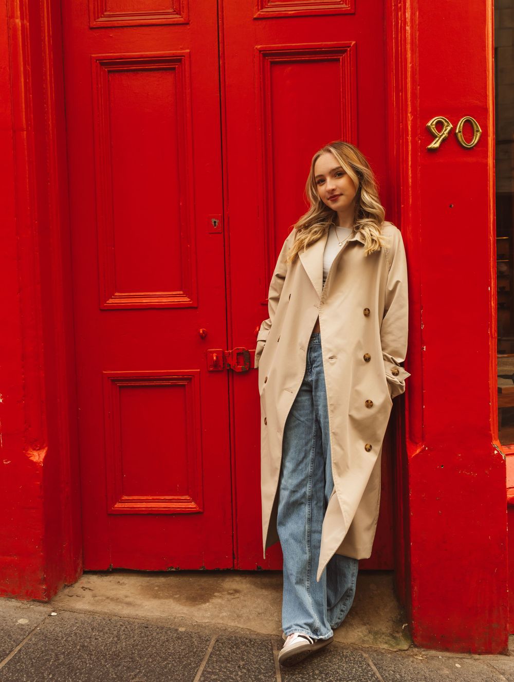 A person poses against vibrant red doors wearing a classic beige trench coat and jeans.