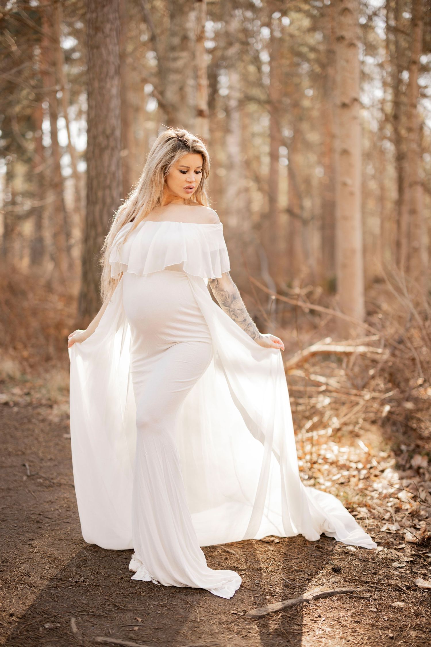 A pregnant woman wearing a flowing white off-shoulder dress poses elegantly in a sunlit forest setting.