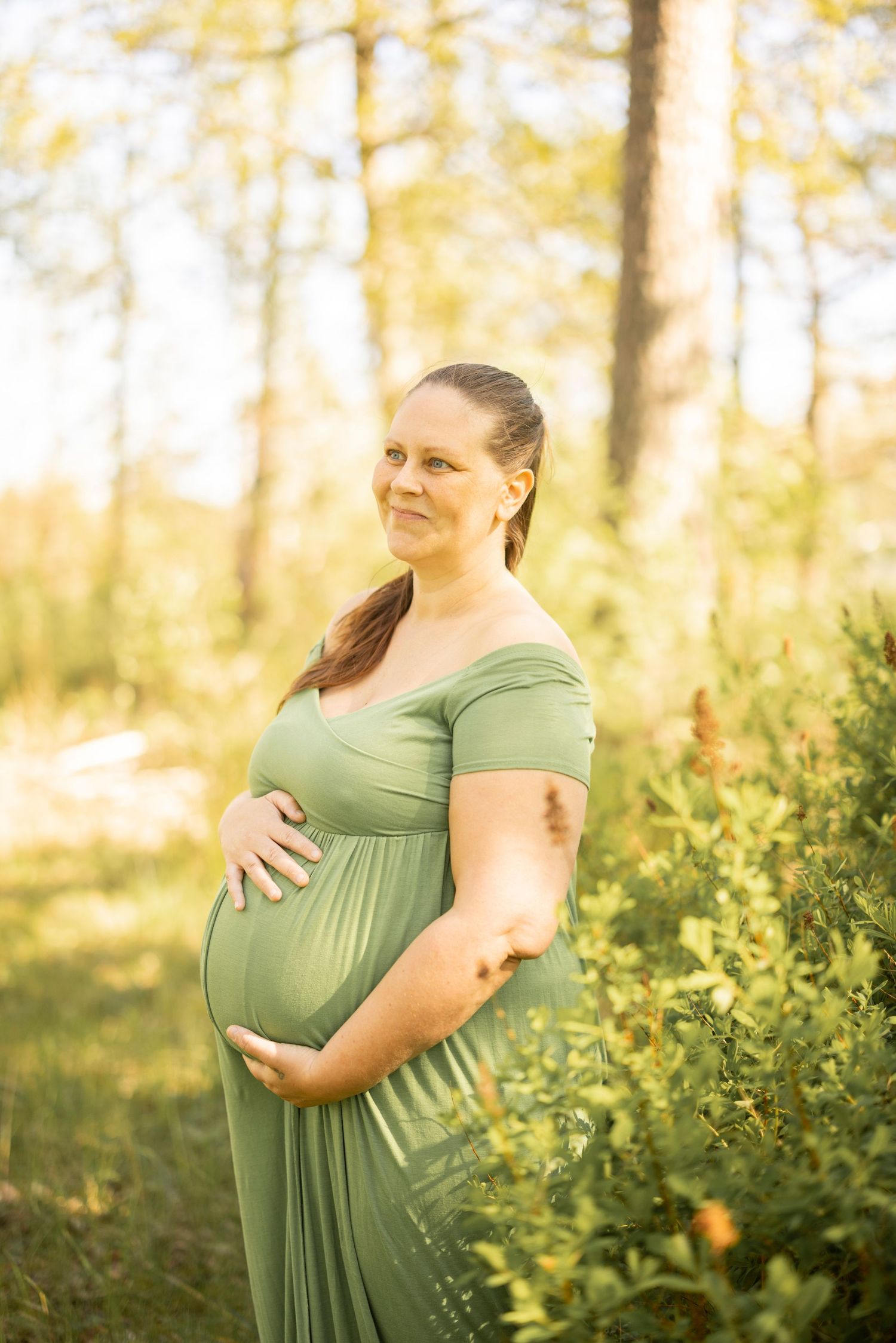 Woman in green dress holds her pregnant belly while standing in nature with golden autumn foliage in background.