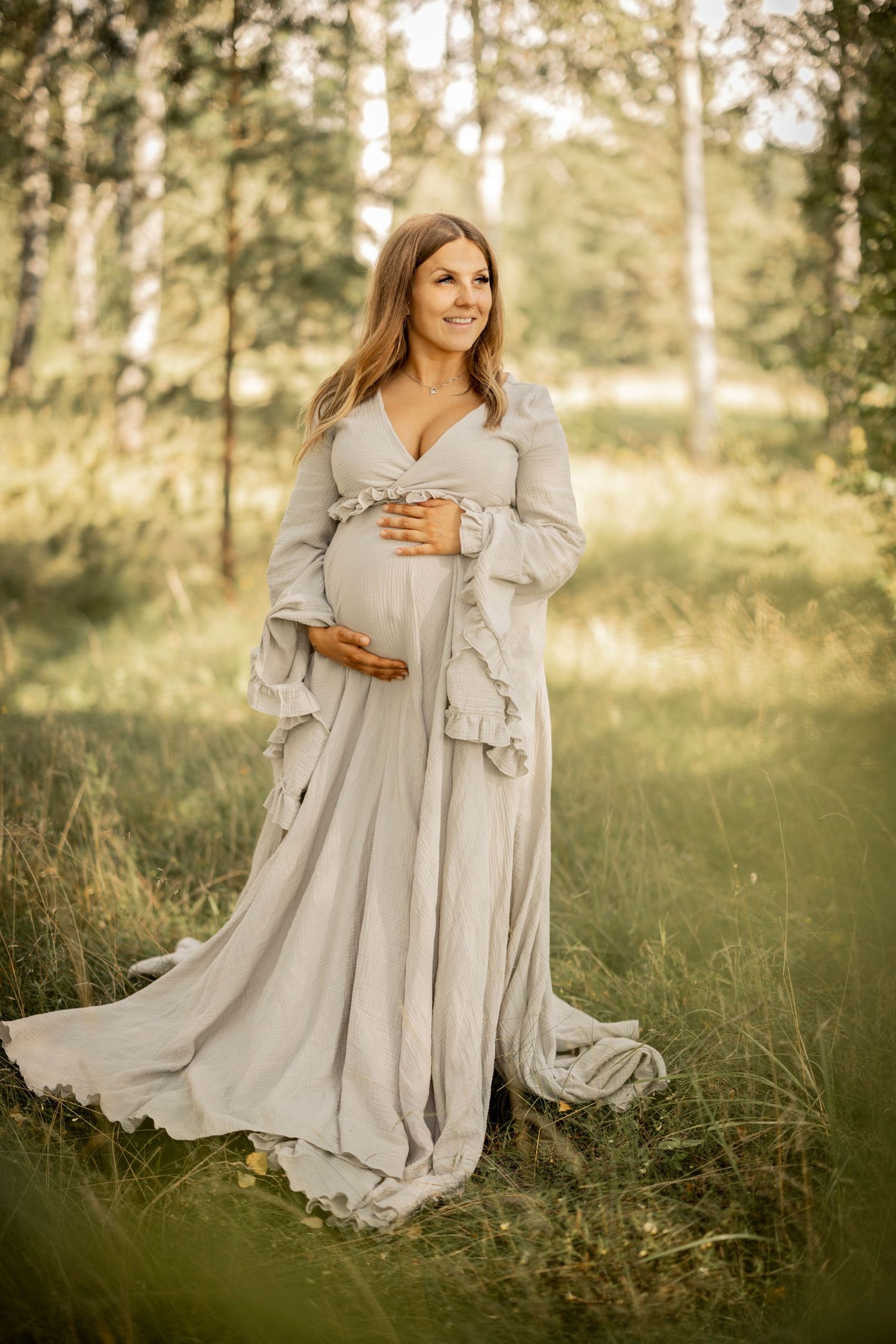 Expectant mother in flowing gray gown stands gracefully in sunlit forest clearing with birch trees in background.