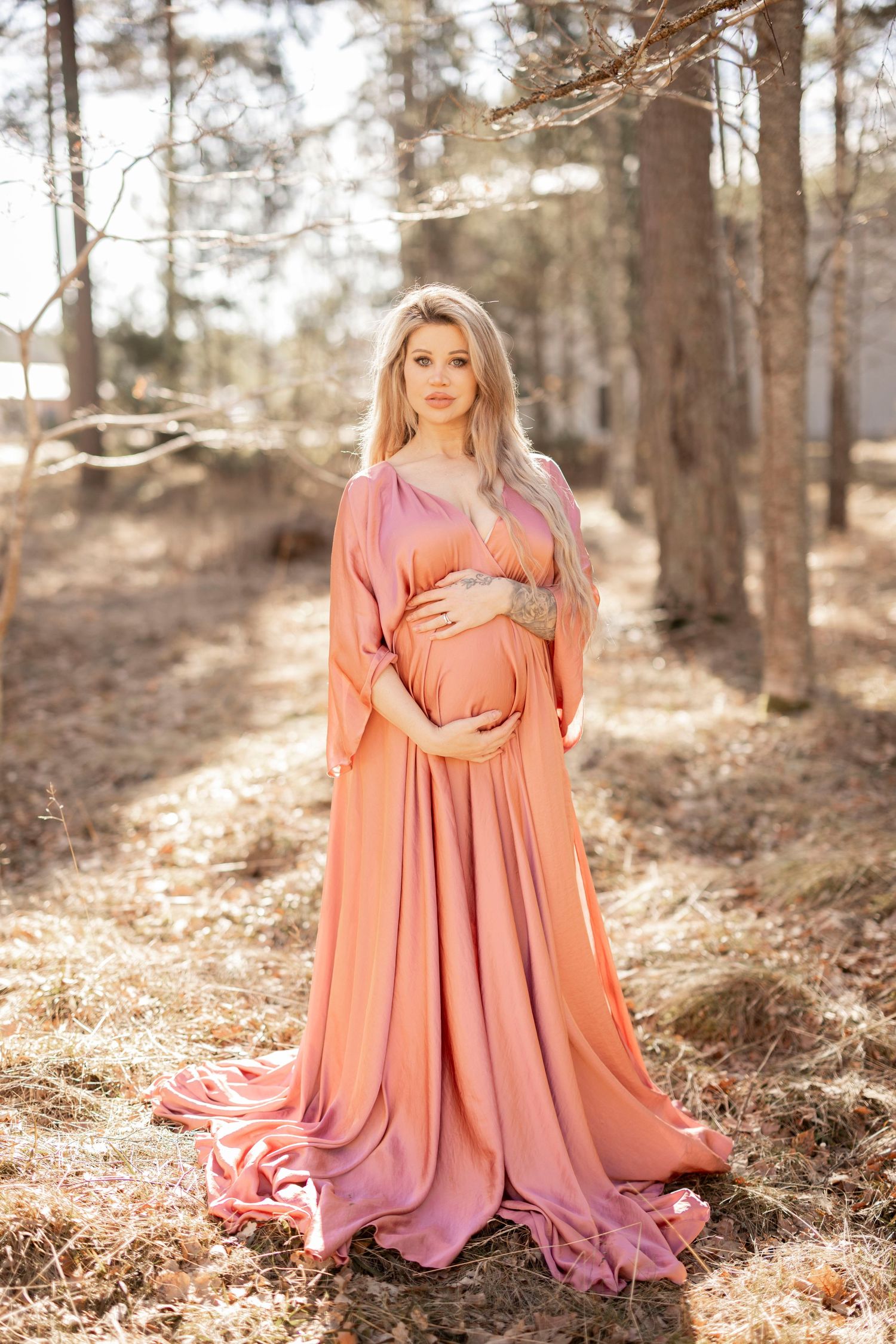 Expectant figure in flowing coral-colored maternity gown poses ethereally in sunlit autumn forest setting.