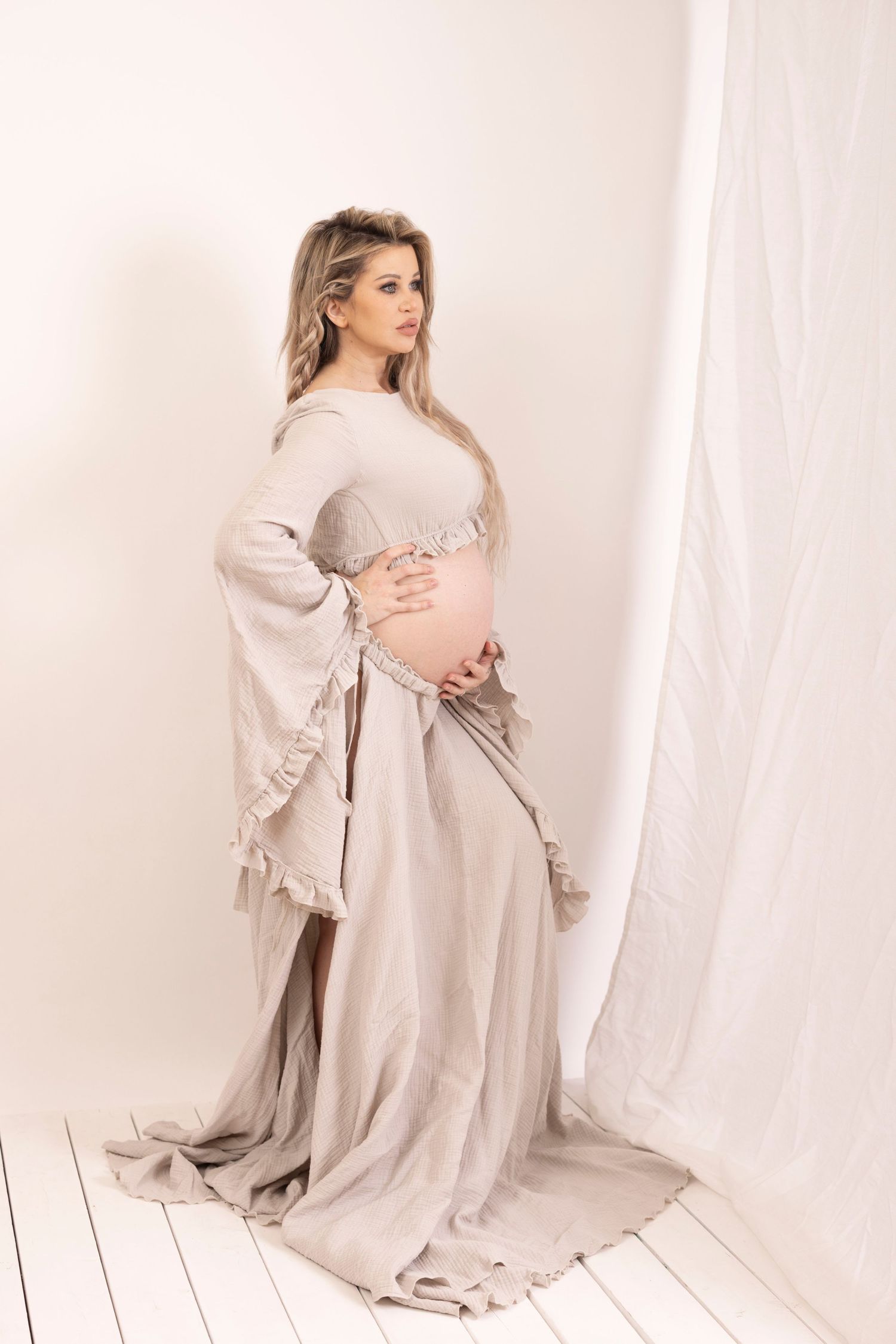 Ethereal maternity photo of an elegant figure in a flowing light gray dress with bell sleeves against white backdrop.