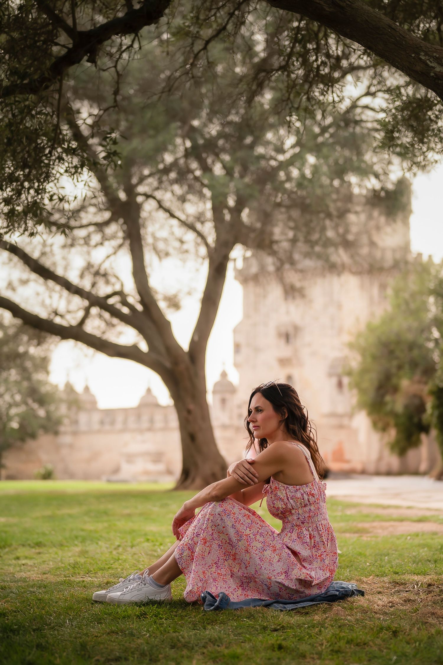 Woman in flowing pink dress sits on grass under large tree near historic stone building.