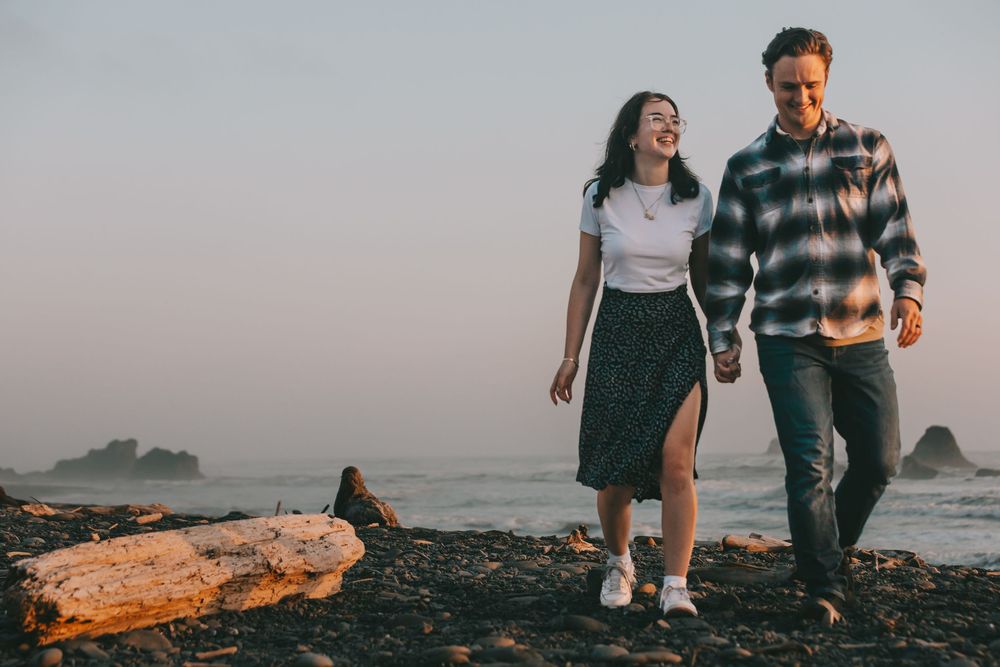 a couple walks along a rocky beach with wood on the beach. She is looking up at him laughing they are holding hands.