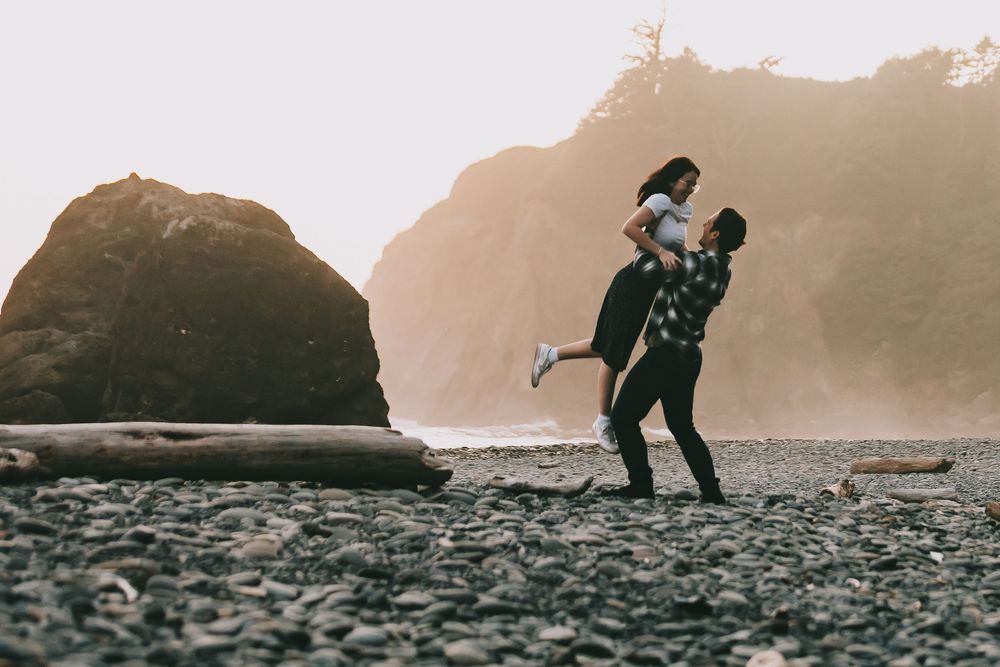 A couple is on a rocky beach with large rocks in the background he is lifting her off the ground and spinning with her.