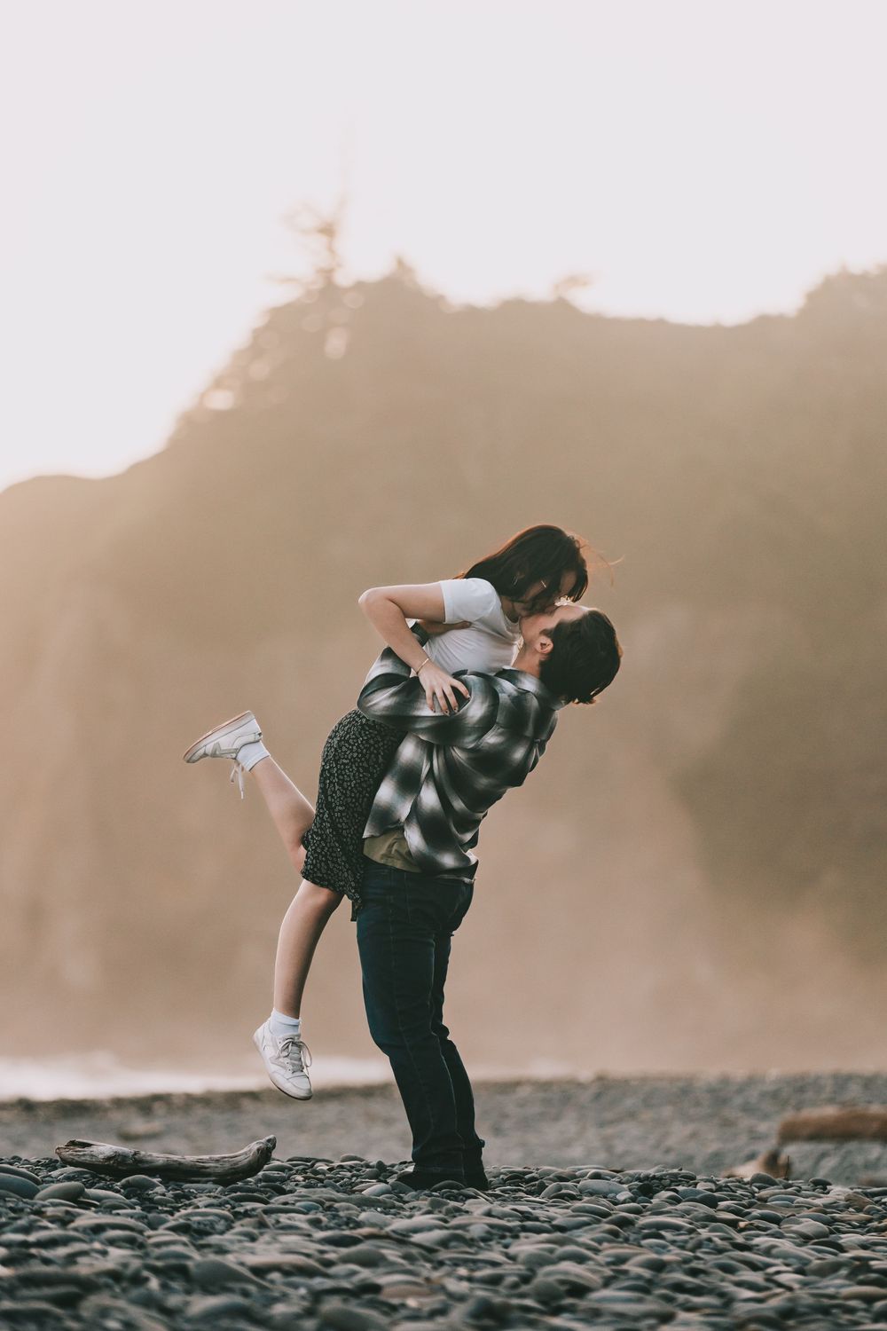 A couple is on a rocky beach with an island in the background he is picking her up to kiss her she has her foot popped.