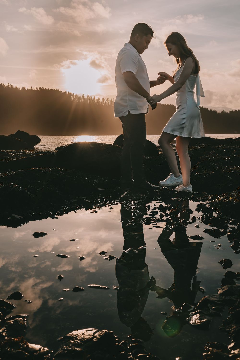 Couple stands on the beach facing each other their reflection in a tide pool. The sun sets behind them.