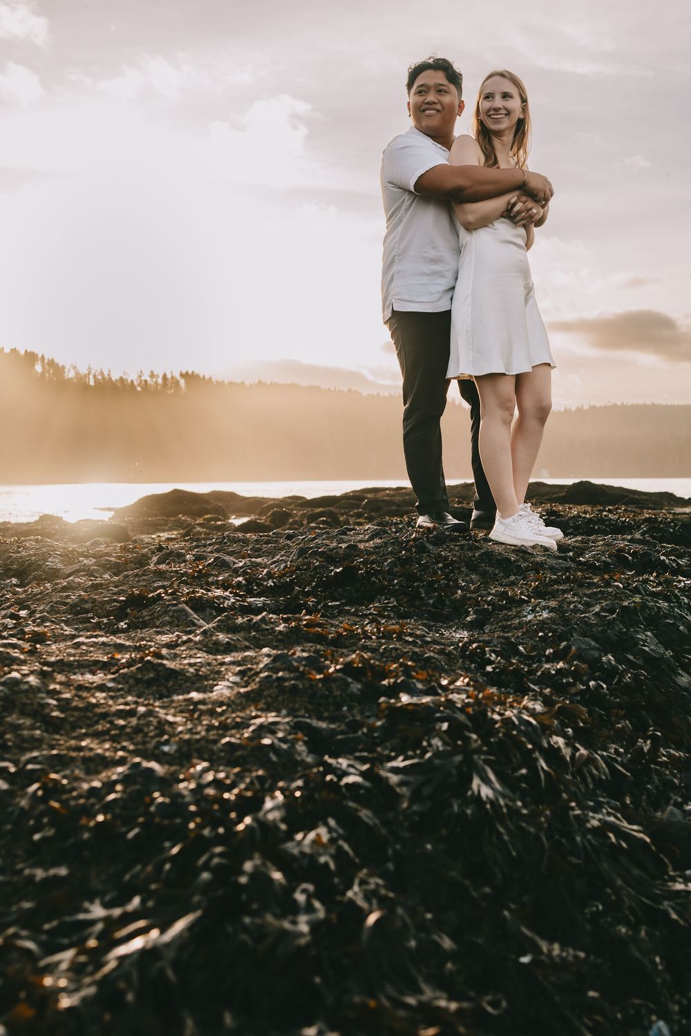 Couple stands on the beach he wraps his arms around her they are both looking off into distance. Sun is setting behind them.