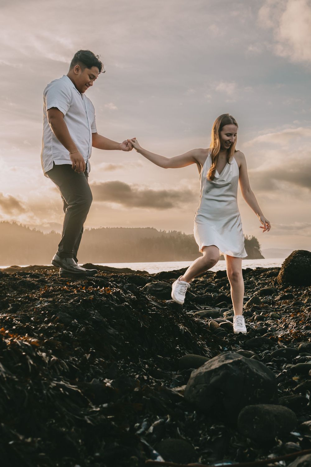 Newly engaged couple walk across rocky beach tide pools during the sunset. She is leading him holding hands