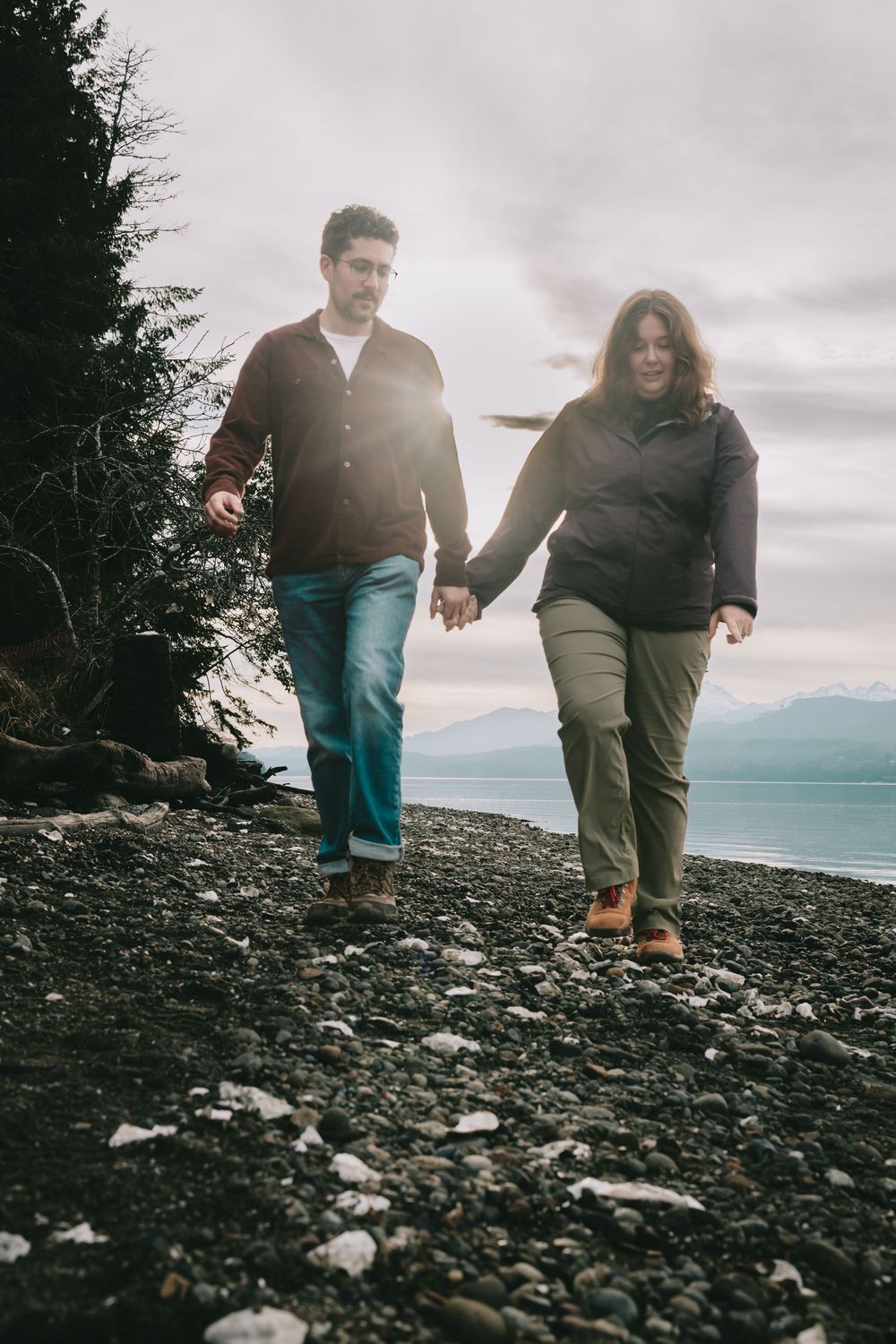 Couple walks along a rocky beach. Beach in the background. Couple is holding hands walking forward.