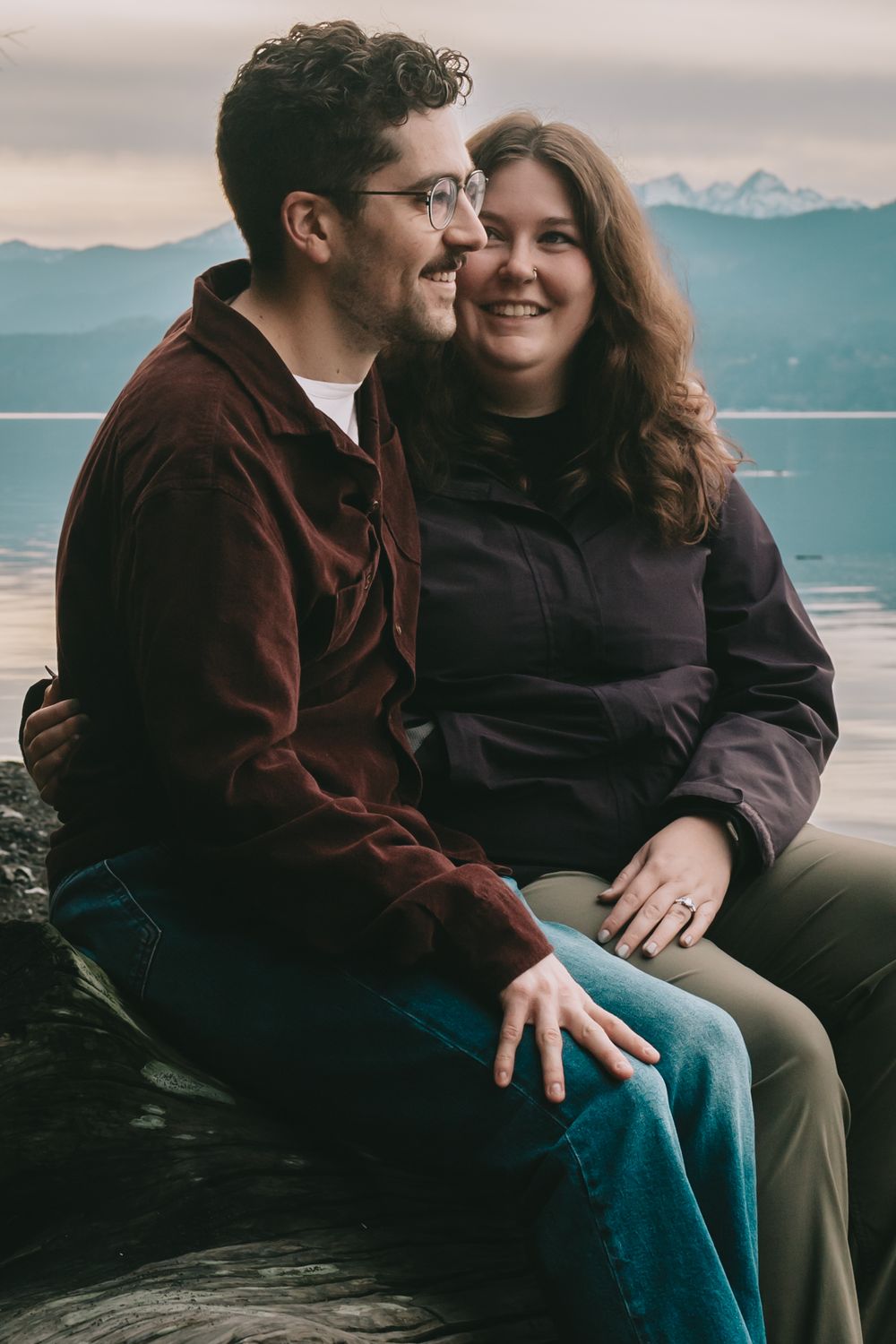 Couple sits on a log on the beach she looks at him he is looking forward. Beach and mountains are behind them.