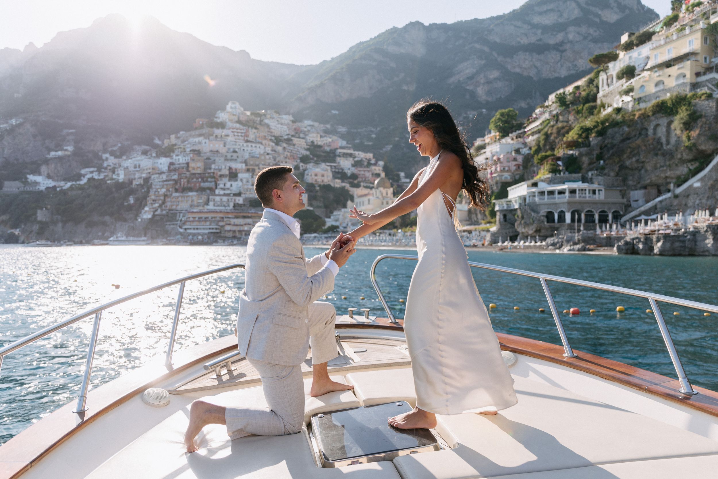 Man proposes to woman on luxury yacht deck with stunning Amalfi Coast cliffside village backdrop on sunny day.