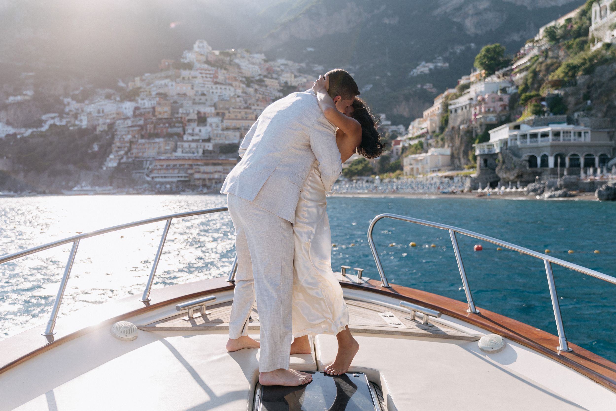 Couple kissing on yacht bow with Positano cliffside village and blue Mediterranean sea in background.