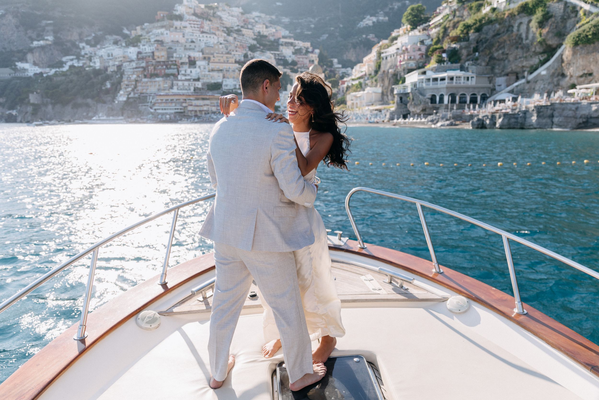Couple in white attire embracing on a luxury yacht bow with Positano coastline in the background.