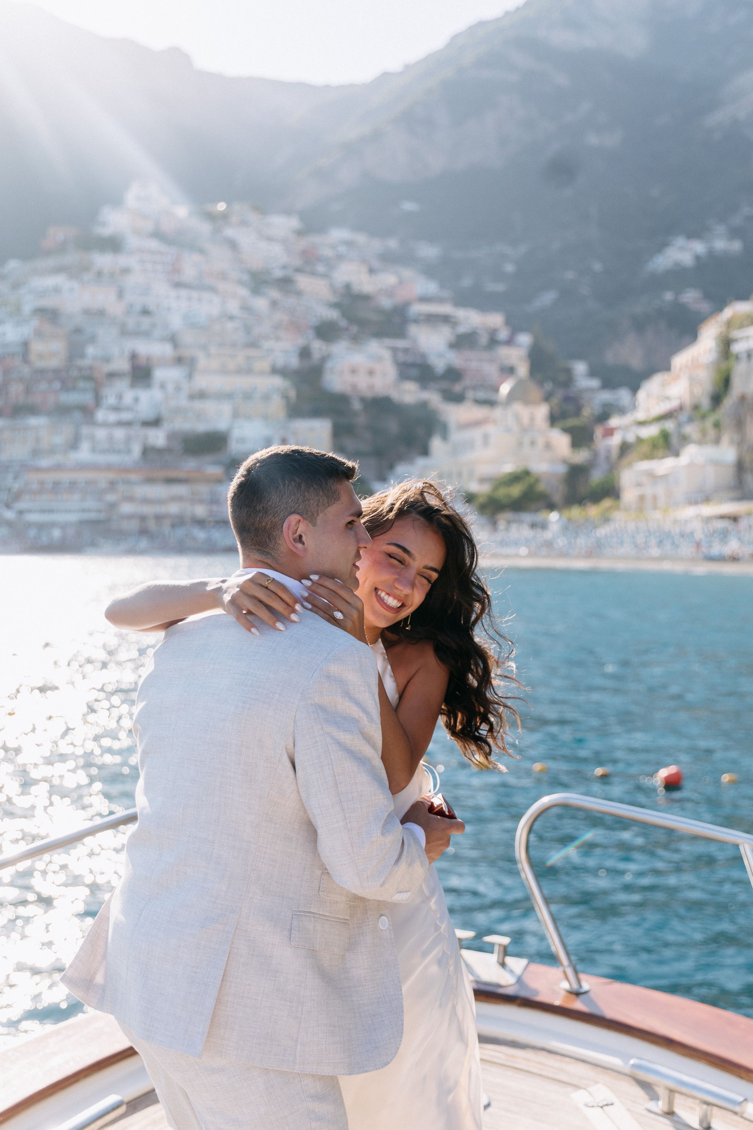 Couple embracing on a yacht with Positano, Amalfi Coast backdrop, sunny day, romantic wedding photo.