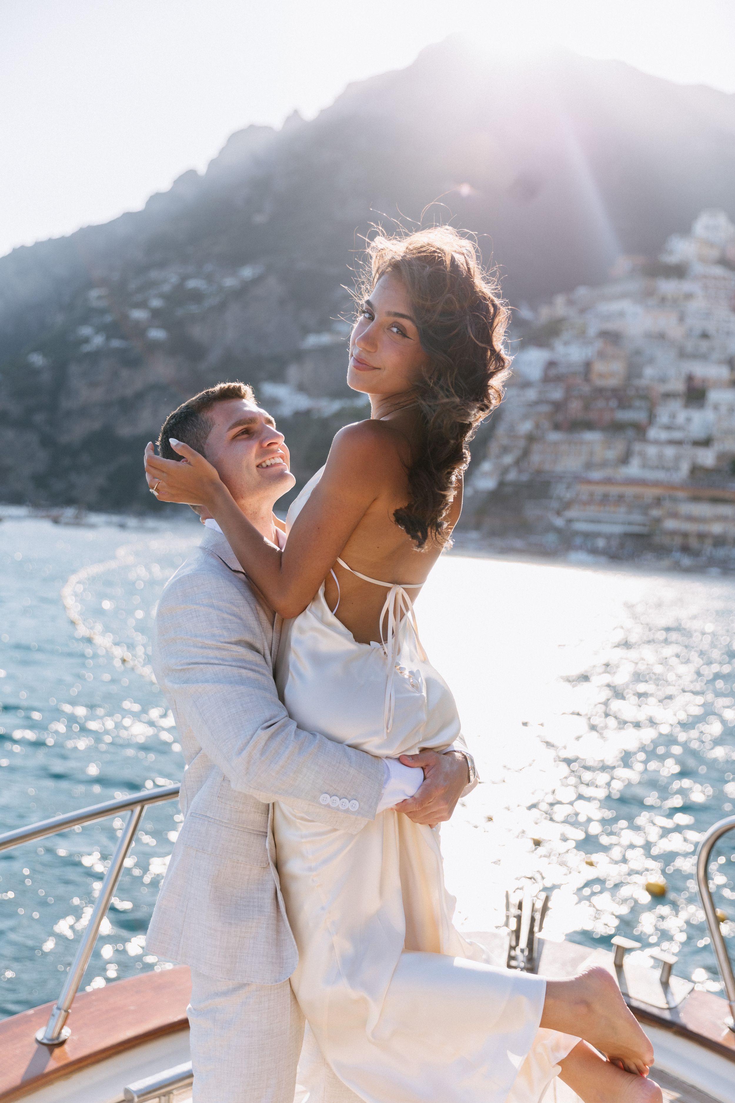 Couple embracing on a yacht at sunset with Positano cliffside village in the background, wearing elegant white outfits.