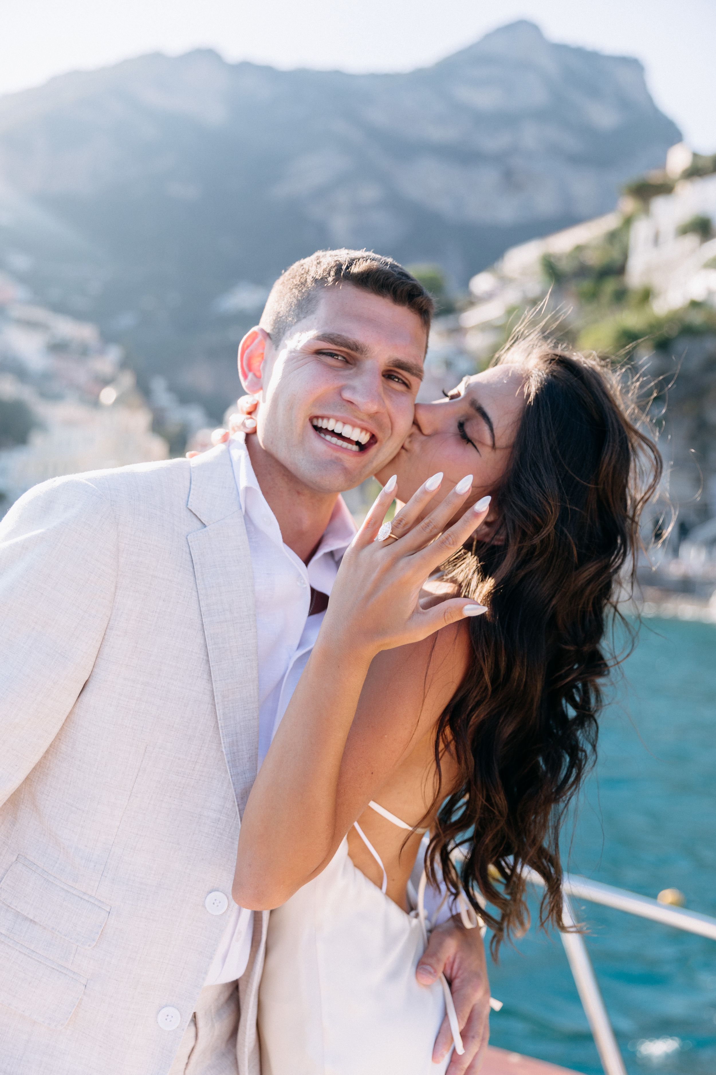 Happy couple embracing on a yacht with stunning Amalfi Coast cliffs, bride showing engagement ring.