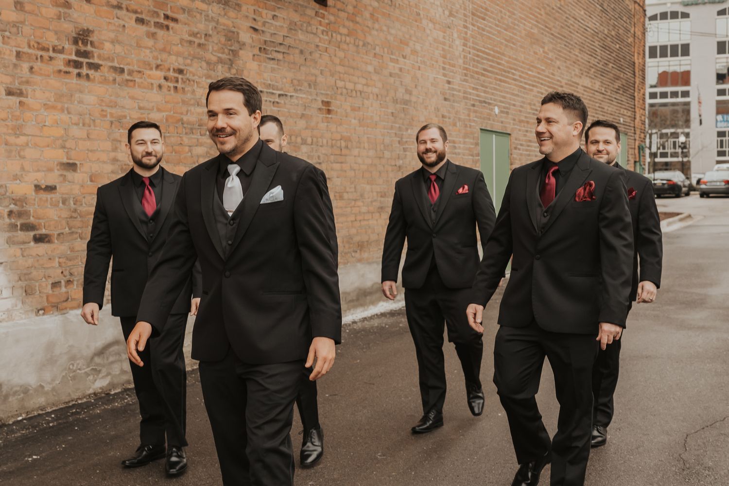 Groomsmen in black suits walk casually along brick wall.