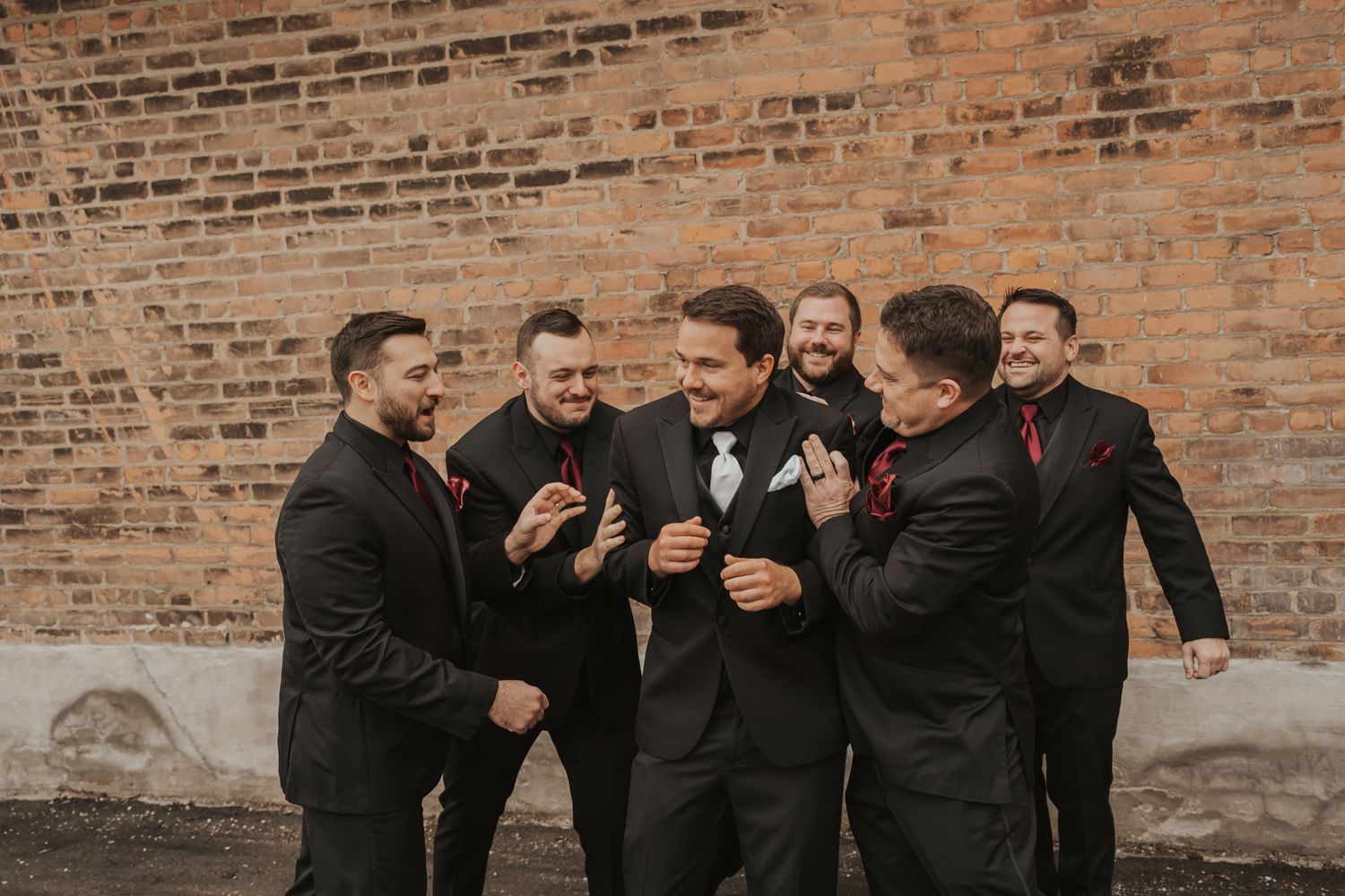 Wedding party members in formal attire gather for photos against brick backdrop.