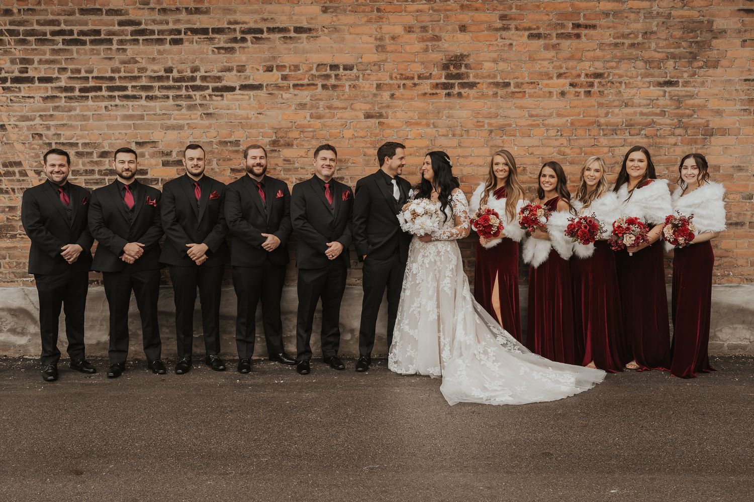 Full wedding party poses together against rustic brick wall background.