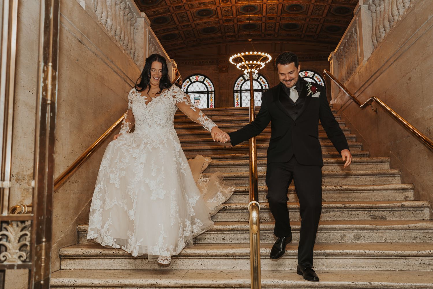 Couple descends grand staircase in historic venue with ornate ceiling.