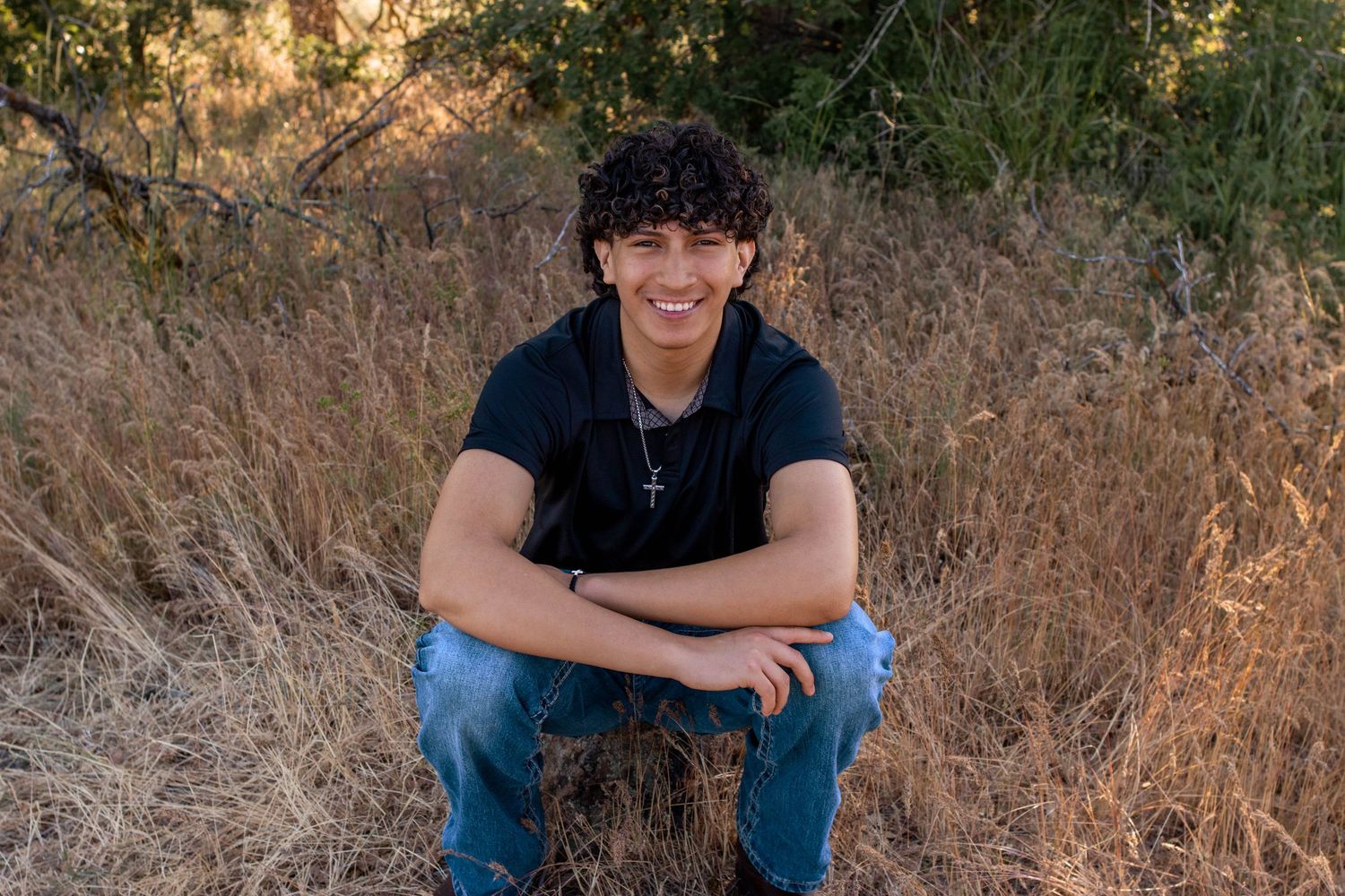 Smiling teenage boy with curly hair sitting cross-legged in a field of golden grass wearing a navy polo shirt.