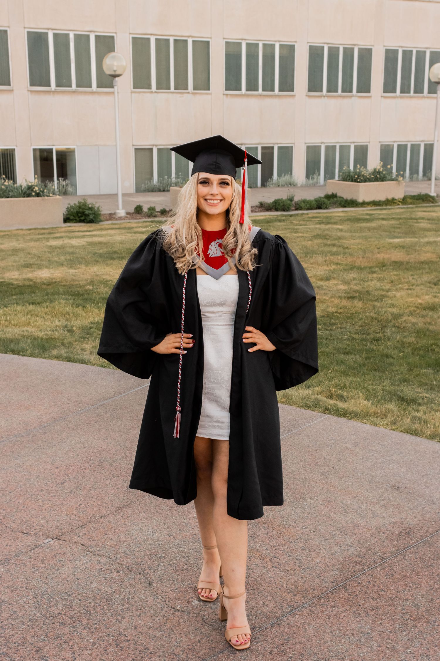 Smiling blonde woman in black graduation cap and gown posing outdoors on campus after commencement ceremony.