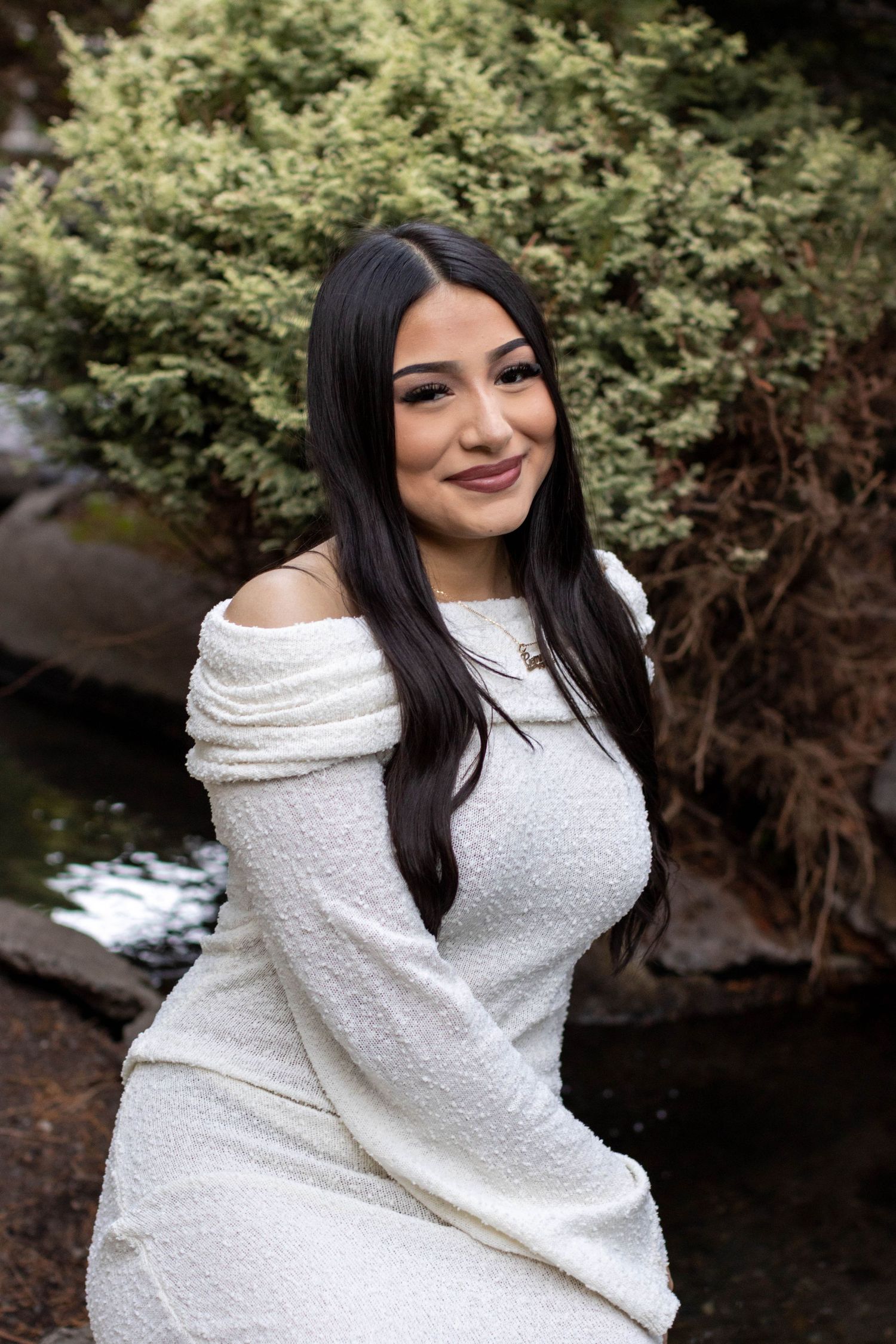 A young woman with long dark hair smiles in a white off-shoulder dress, posed near rocks and greenery outdoors.
