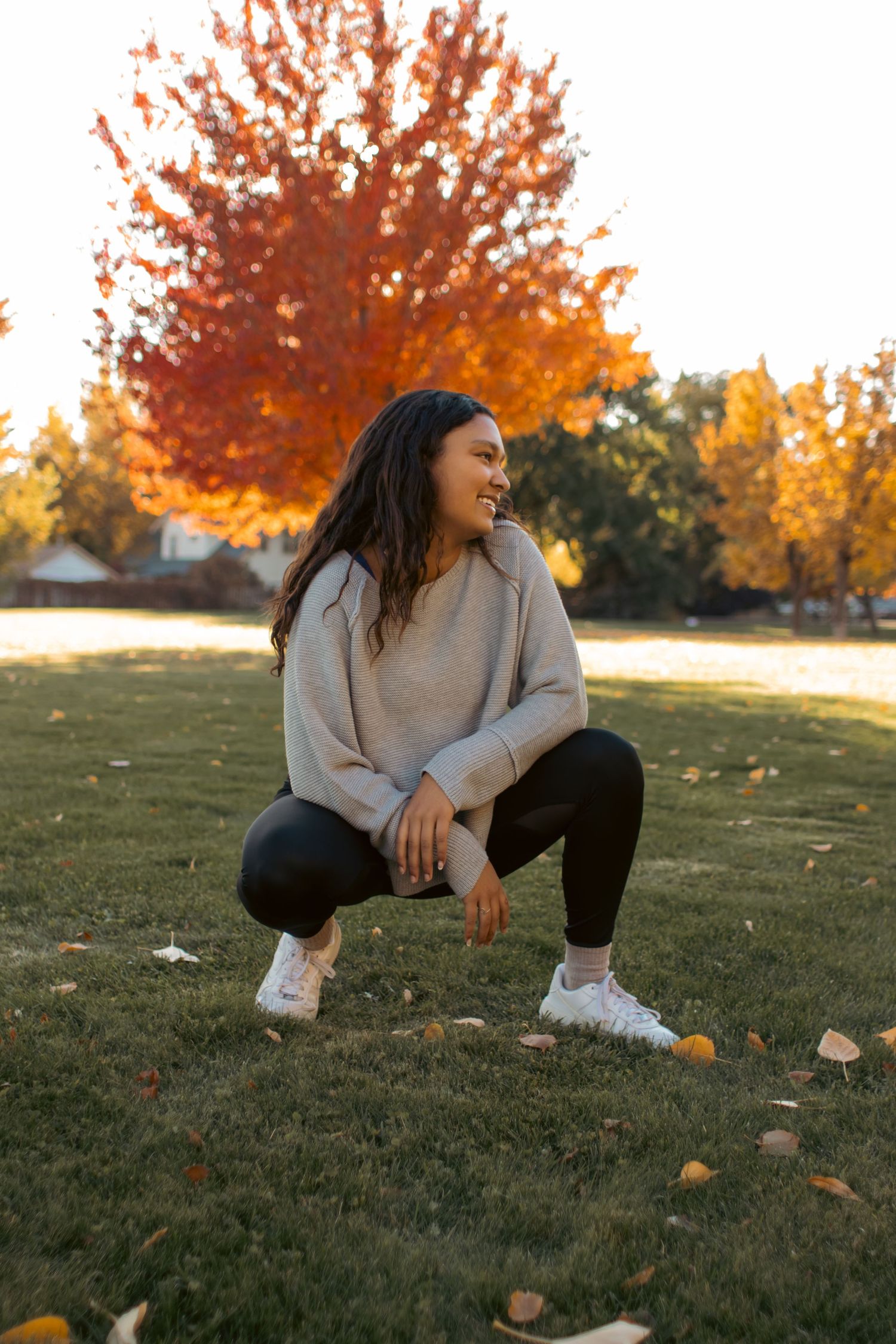 Woman crouching on grass in autumn park, wearing beige sweater and black leggings, smiling with colorful fall foliage behind her.