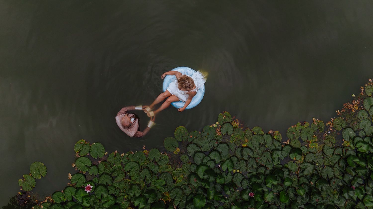 Luchtfoto van een bruid in het wit die op een luchtmatras in een meer ligt, omringd door waterlelies.