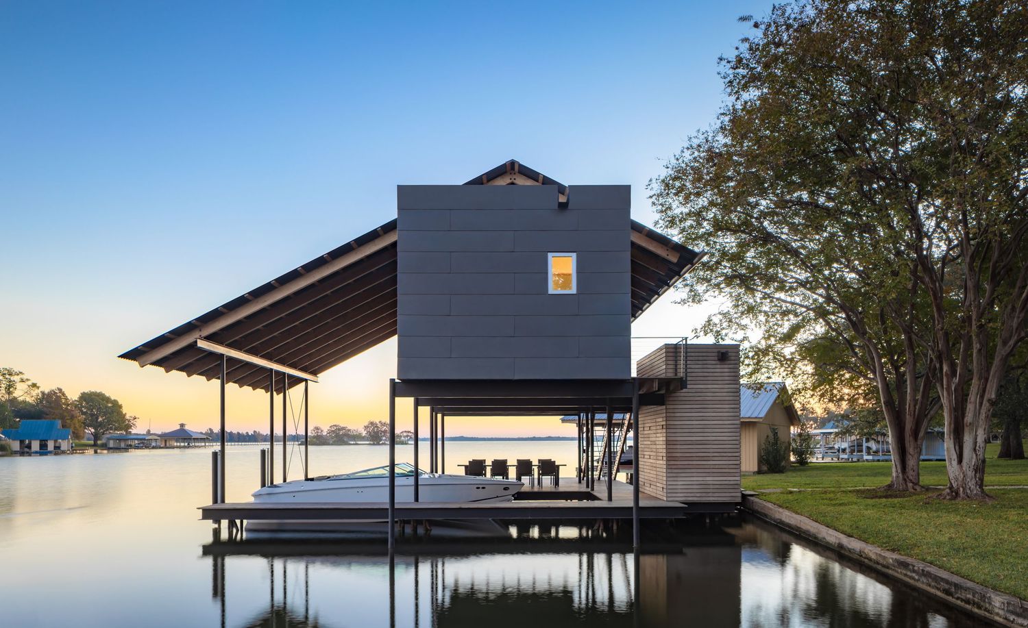 Modern boathouse on stilts over calm lake water at sunset, featuring dark metal cladding and open pavilion design.