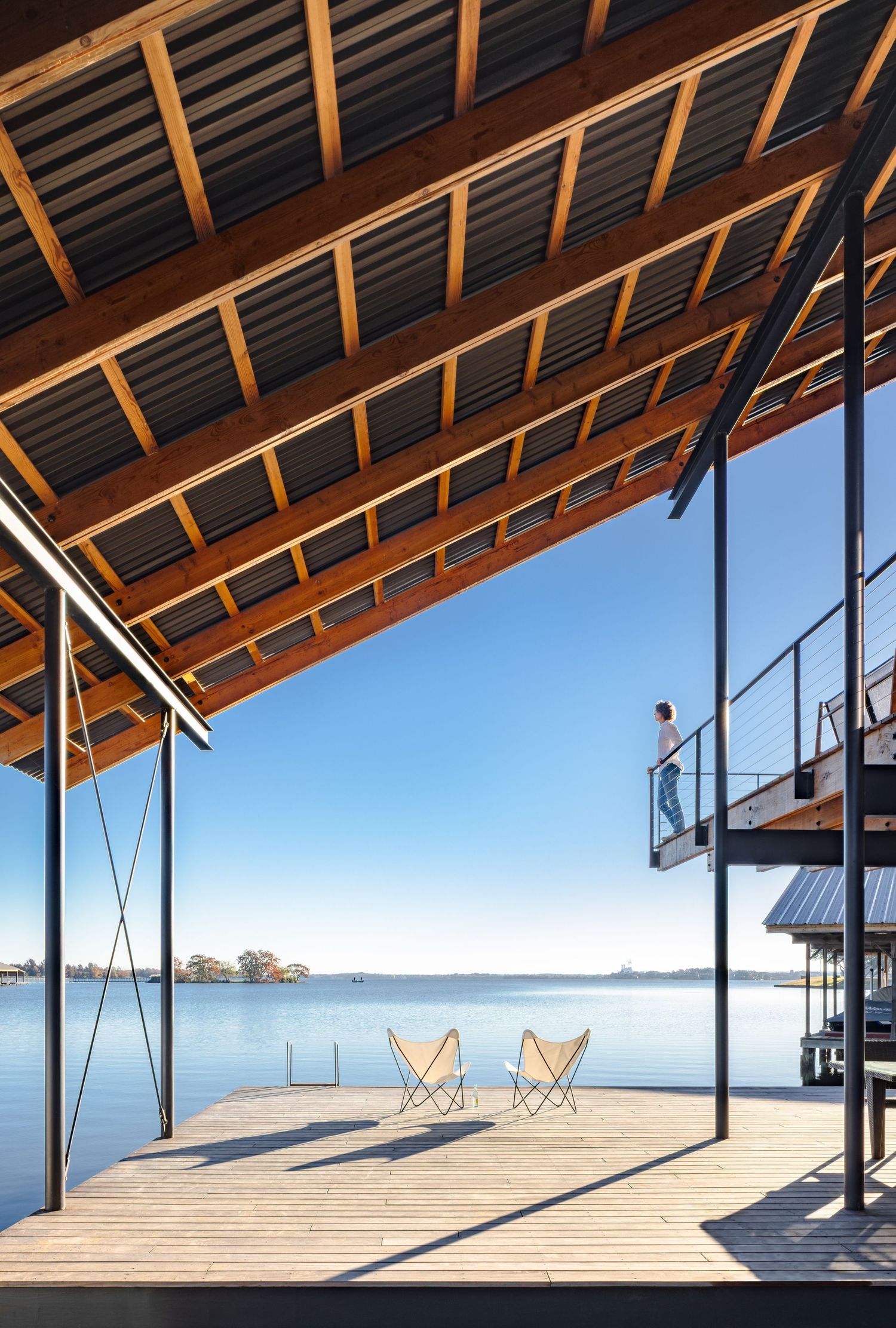 Modern lakeside deck with butterfly chairs under a wooden roof, overlooking calm blue water on a sunny day.