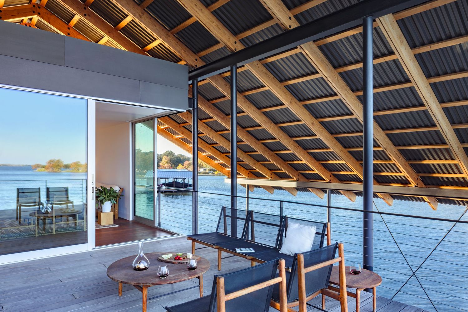 Modern overwater deck with teak furniture, corrugated roof, and panoramic ocean views through floor-to-ceiling glass walls.