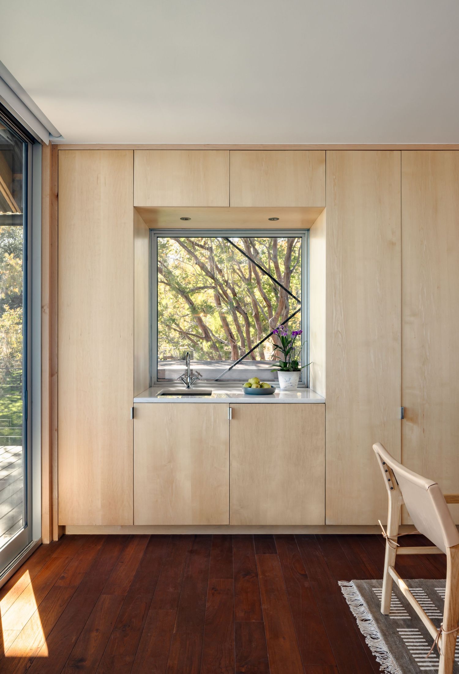 Modern kitchen nook with light wood cabinetry, dark hardwood floors, and a framed window overlooking lush green trees.