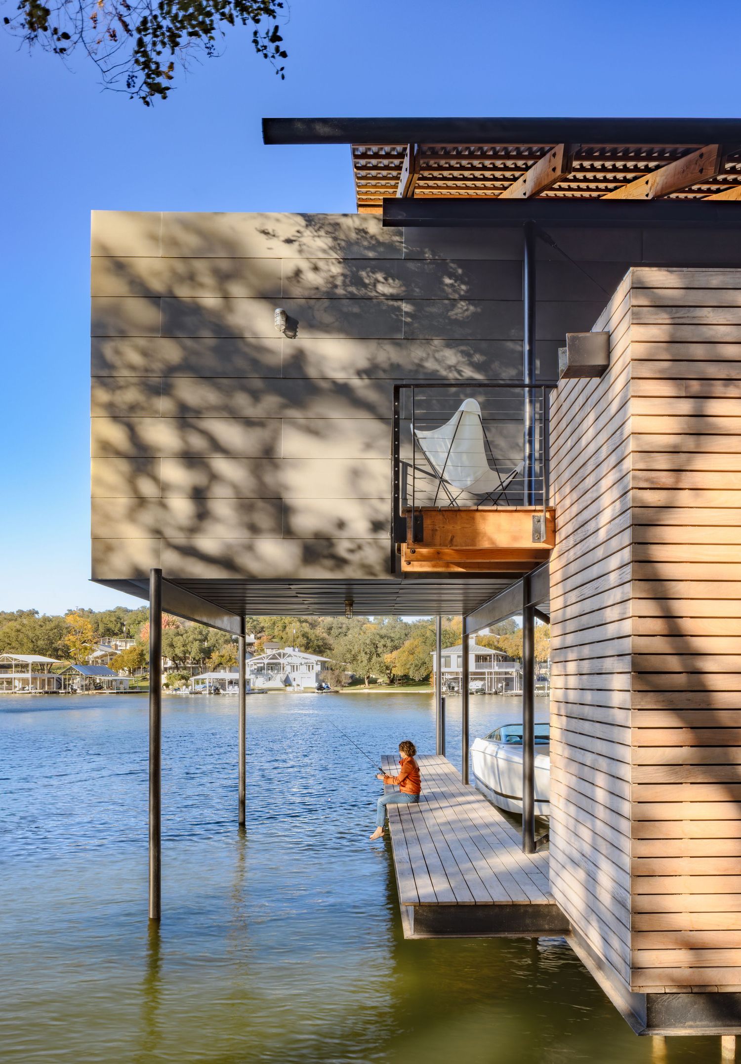 Modern lakeside boathouse on stilts with wood cladding, featuring a dock where a child fishes over calm blue water.