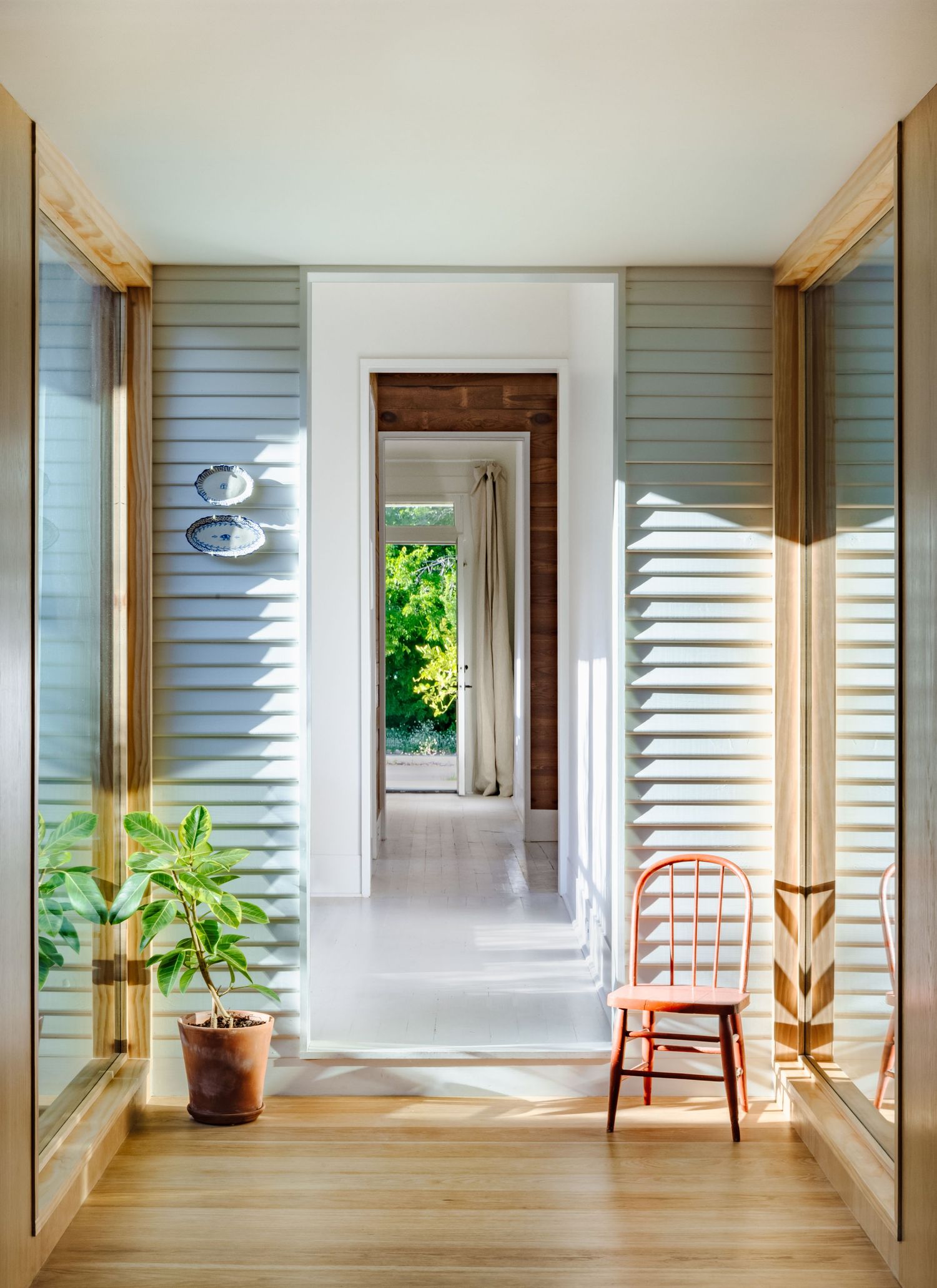 Bright hallway with slatted walls, wooden floors, and a single orange chair near large windows.