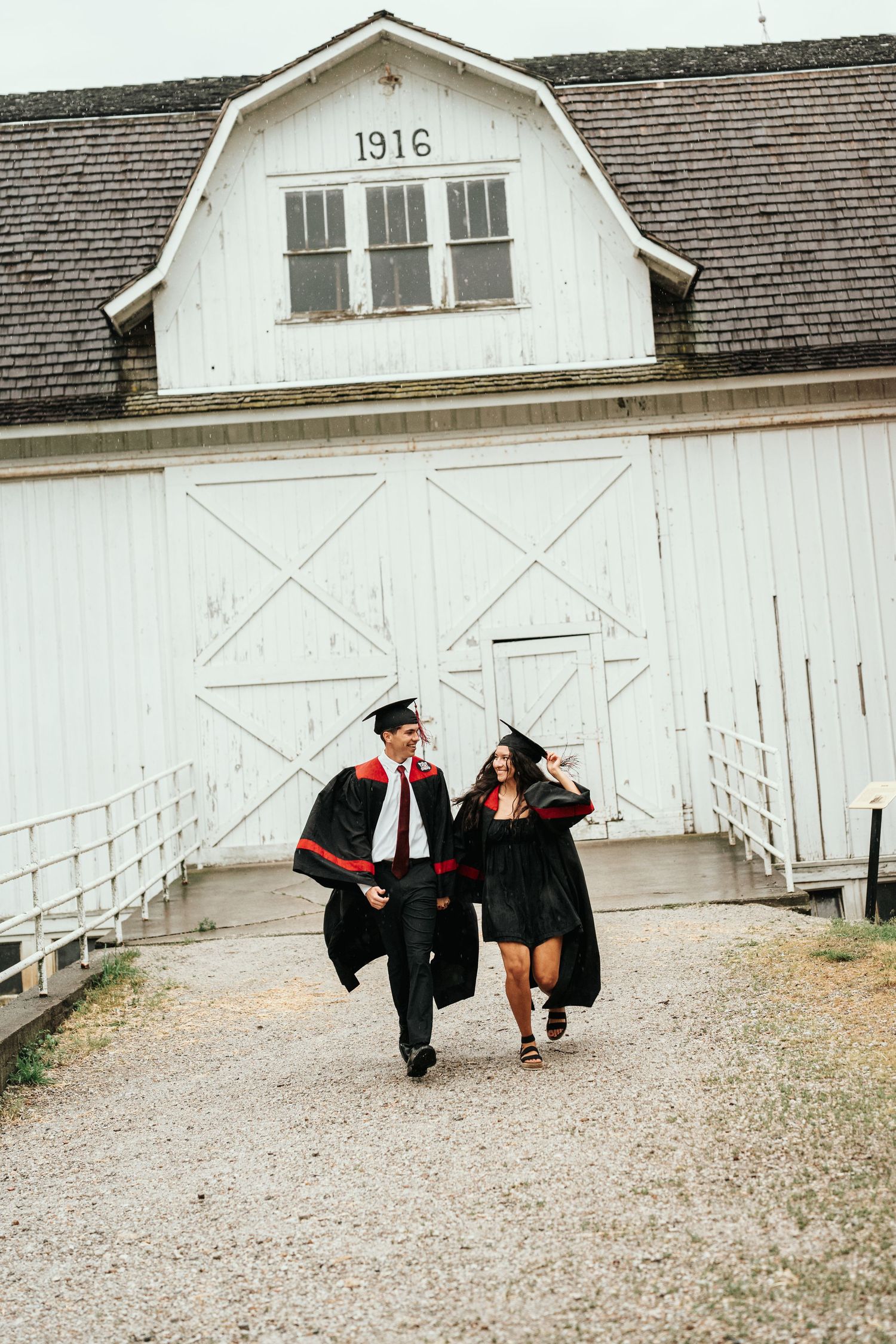 Two graduates in black caps and gowns walk together in front of a white barn dated 1916.