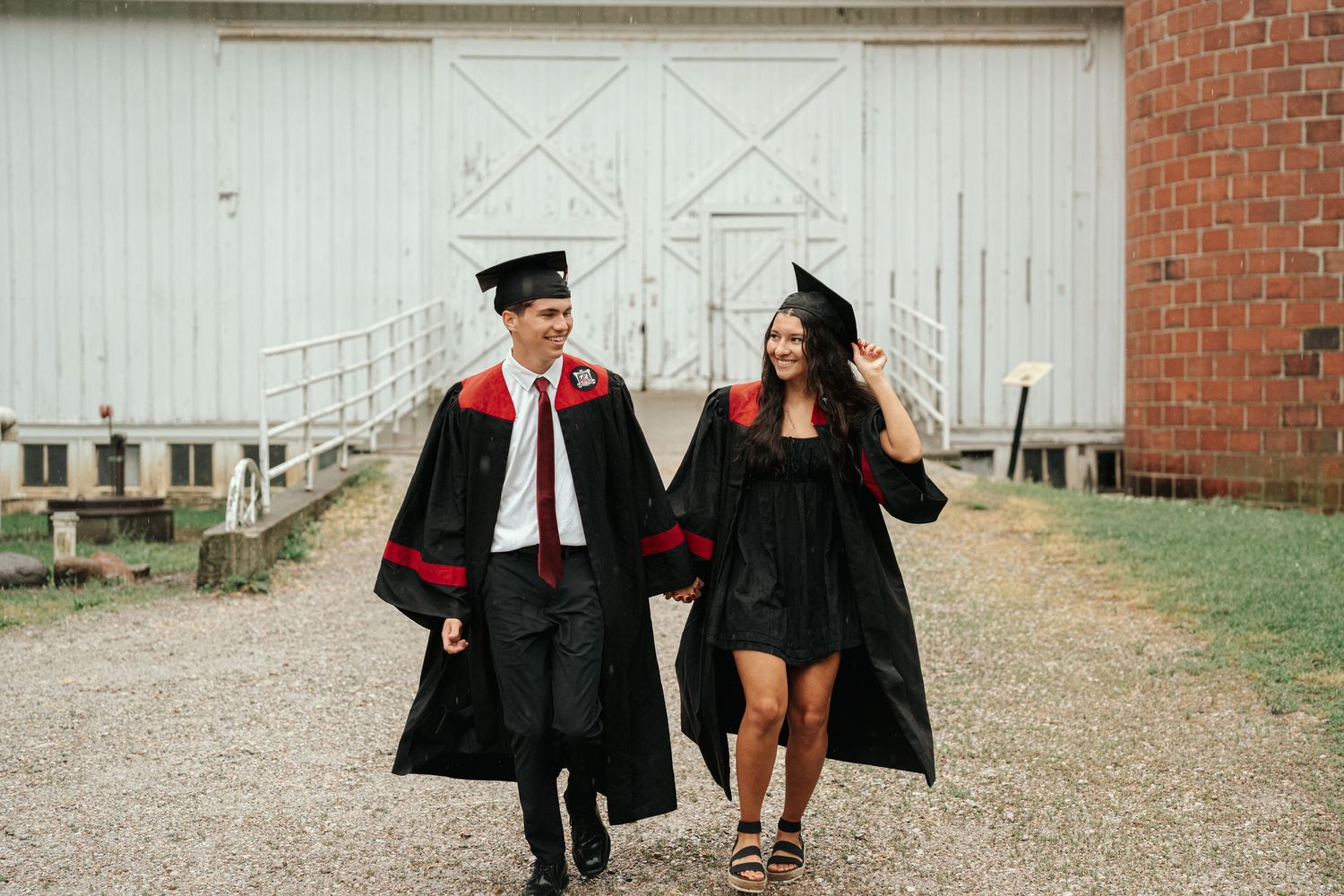 Two graduates in black and red caps and gowns walking together outside a white barn.